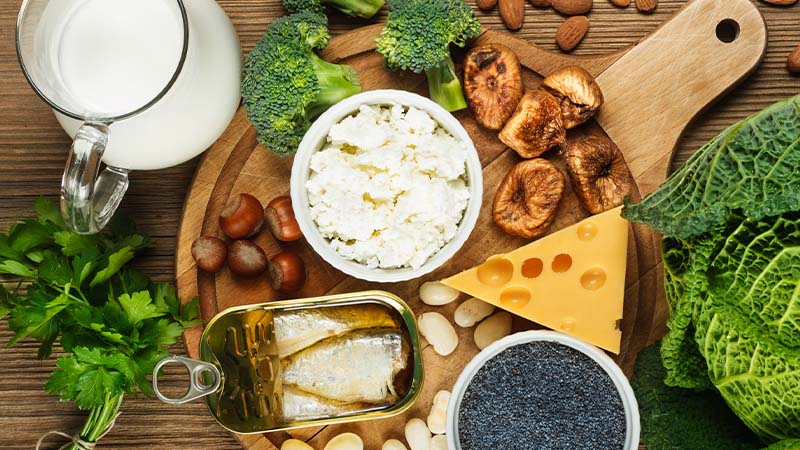 Mesa com alimentos ricos em cálcio: leite, queijo, iogurte, frutos secos, sementes, azeite, brócolos e folhas verdes, dispostos sobre madeira